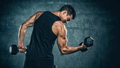 Ein Mann im Tank Top steht mit dem Rücken zur Kamera und hält zwei Kurzhanteln in den Händen. Er dreht sich leicht nach rechts und spannt seinen rechten Arm an.