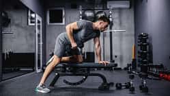 Ein muskulöser Mann trainiert mit einer Hantel, die sich auf einer Sportbank im Fitnessraum befindet. 