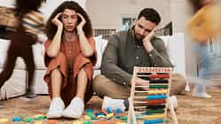 Eine Mutter und ein Vater sitzen verzweifelt auf dem Boden des Kinderzimmers, ihre beiden Kinder toben um die beiden herum