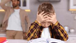 Ein kleiner Junge sitzt angestrengt über einem Schulbuch, im Hintergrund sieht man einen verzweifelten Vater