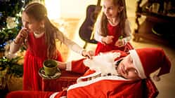 Ein als Weihnachtsmann verkleideter Vater sitzt schlafend in einem Stuhl, während ihm seine beiden Töchter den falschen Bart vom Gesicht ziehen