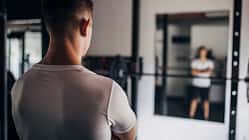 Mann steht mit dem Rücken zum Betrachter in einem leeren Fitnessstudio vor dem Spiegel