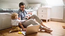Ein Vater sitzt vor einem Sofa, mit einem aufgeklappten Notebook und zwei kleinen Kindern an seiner Seite
