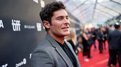 Zac Efron steht in grauem Anzug bei einer Filpremiere vor einer Werbewand.