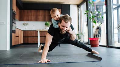 Ein Vater macht Liegestütze in der Küche während sich sein Baby auf seinem Rücken festhält