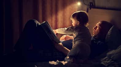 Ein Vater liegt mit seinem Kind im Bett und liest ihm aus einem Buch vor