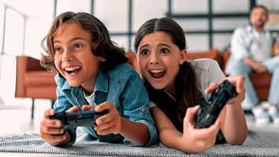 Man sieht zwei Kinder auf dem Teppich liegend auf einer Konsole spielen. Im Hintergrund sitzt der Vater auf dem Sofa.