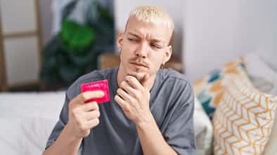 Young,Caucasian,Man,Holding,Condom,Sitting,On,Bed,Serious,Face