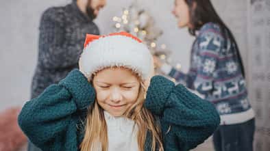 Ein Mädchen mit Weihnachtsmütze hält sich die Ohren zu, im Hintergrund sieht man, wie sich die Eltern lautstark streiten