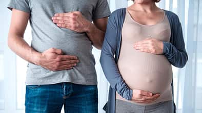 Eine schwangere Frau hält sich den Bauch, daneben steht ein Mann mit einem kleinen Bauchansatz in der selben Körperhaltung