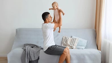 Ein Vater sitzt auf dem Sofa, hält sein Baby in die Höhe und riecht an der Windel