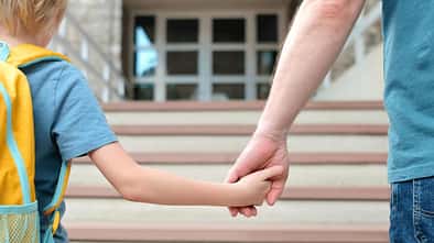 Man sieht in einer Nahaufnahme Vater und Sohn von hinten Hand in Hand zur Schule gehen.