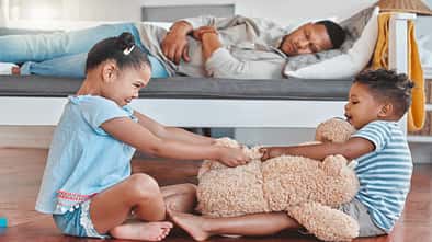 Zwei Kinder streiten sich im Wohnzimmer, dahinter liegt der Vater erschöpft auf einer Couch
