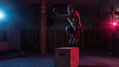 Muscular man trains jumping on a wooden box in a dark gym. Athlete Workout in red blue neon light. Cross, functional training