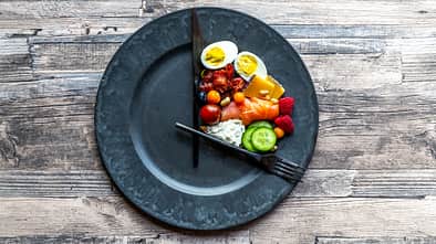 verschiedenes gesundes Essen auf einem runden Teller mit Besteck als Uhrzeiger