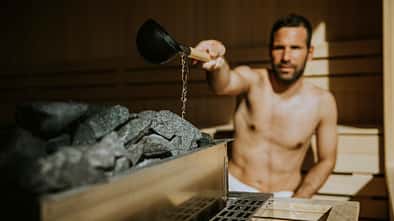 Junger Mann, der in der Sauna Wasser auf heißen Stein gießt