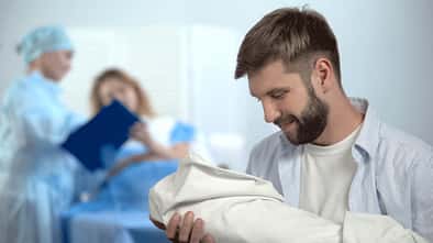 Ein Vater hält sein neugeborenes Kind im Arm, im Hintergrund sieht man die Wöchnerin im Bett liegen und eine Krankenschwester.