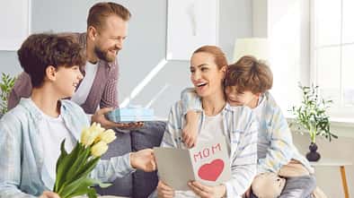 Eine Frau sitzt auf dem Sofa, ihr Mann und ihre Kinder reichen ihr Blumen und Schokolade und eine Karte zum Muttertag