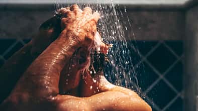 GettyImages.de/Mariia Siurtukova Engrossed in his grooming ritual, a man rinses his hair, the water's embrace enhancing the meditative quality of his daily routine.