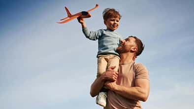 Ein Vater hat seinen kleinen Sohn auf dem Arm, der einen Papierflieger in den Himmel starten lässt