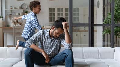 Auf dem Sofa sitzt ein verzweifelter Vater, dahinter tobt sein Sohn herum