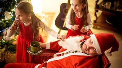 Ein als Weihnachtsmann verkleideter Vater sitzt schlafend in einem Stuhl, während ihm seine beiden Töchter den falschen Bart vom Gesicht ziehen