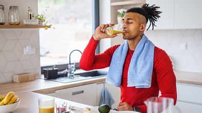 Sportlicher Mann mit Handtuch um den Hals trinkt ein Glas Orangensaft in einer hellen Küche, auf der Arbeitsfläche liegt Obst und Gemüse