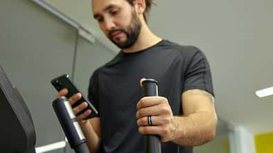 Ein Mann trainiert auf einem Crosstrainer im Fitnessstudio, hält einen Griff des Geräts und blickt auf sein Smartphone. Er trägt einen Fitnessring