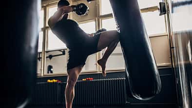 Mann trainiert Kampfsport im Boxstudio und tritt mit einem Bein gegen einen Boxsack.