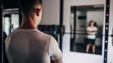 Mann steht mit dem Rücken zum Betrachter in einem leeren Fitnessstudio vor dem Spiegel