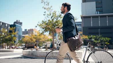 Geschäftsmann schiebt mit Kaffeebecher und Umhängetasche sein Fahrrad durch die Stadt