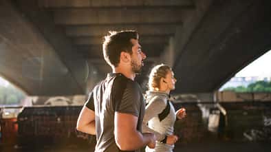 Mann und Frau joggen unter einer Brücke durch, die Sonne im Rücken
