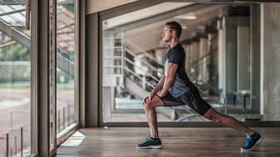 Sportlicher Mann macht einen Ausfallschritt-Stretch in einer modernen Trainingshalle vor großen Fenstern.