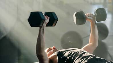 Mann liegt auf einer Trainingsbank und führt im Fitnessstudio eine Kurzhantel-Bankdrück-Übung aus.