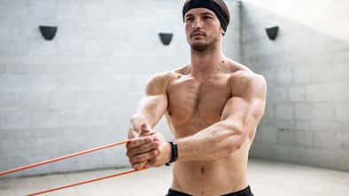 Ein Mann trainiert oberkörperfrei in einem Raum und hält mit beiden Armen ein Stretching-Band