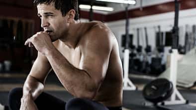 Sportlicher Mann in Sportleggings und oberkörperfrei sitzt im Fitnessstudio in der Hocke und sieht erschöpft aus