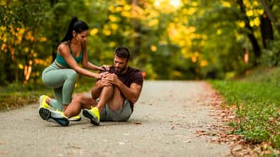 Ein Mann hält sich beim Joggen in der Natur das Knie, während er auf dem Boden sitzt und eine Frau kümmert sich um ihn