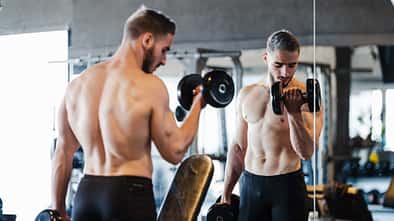 Junger muskulöser Mann macht Kurzhantel-Übungen im Fitnessstudio vor einem Spiegel