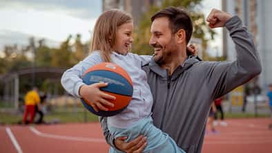 Ein Mann, der einen Ball und seine Tochter im Arm hält und seinen Bizeps anspannt Ein Mann, der einen Ball und seine Tochter im Arm hält und seinen Bizeps anspannt