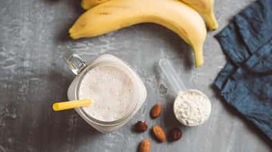 Proteinsmoothie im Glas mit Mandeln, Banane und einem Scoop Proteinpulver daneben