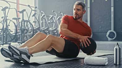 Mann führt eine Bauch-Übung mit einem Ball auf einet Sportmatte aus, im Hintergrund sind Sportgeräte zu sehen
