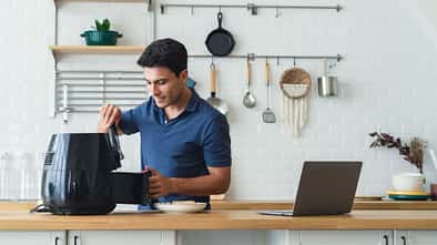 Ein Mann steht in der Küche, neben ihm ein Laptop, und bereitet Essen im Airfryer zu