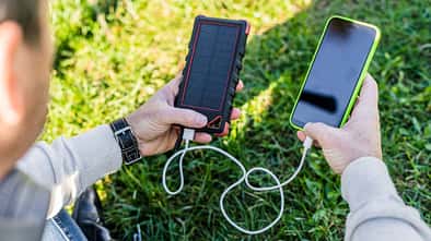 Person sitzt auf einer Wiese und hält eine Solar-Powerbank in der einen und ein Smartphone, welches aufgeladen wird, in der anderen Hand