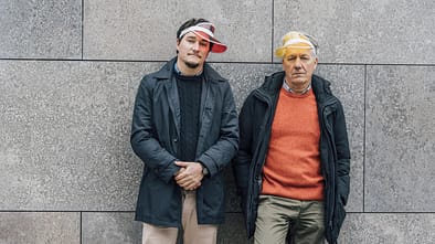Ein älterer und ein jüngerer Mann stehen vor einer Mauer und tragen beide eine ähnliche Schirmmütze