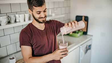 Junger Mann steht in der Küche und schenkt sich einen frisch gemixten Proteinshake in ein Glas ein