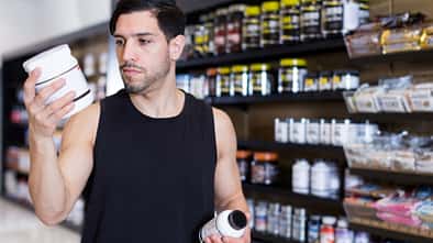Ein Mann steht vor einem Regal mit Protein-Supplementen und schaut sich die Inhaltsstoffe von zwei Produkten an