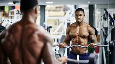 Ein muskulöser Mann trainiert mit einer Langhantel vor einem Spiegel im Fitnessstudio