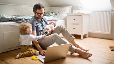 Ein Vater sitzt vor einem Sofa, mit einem aufgeklappten Notebook und zwei kleinen Kindern an seiner Seite