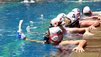 unterwasserhockey Mehrere Schwimmer:innen in Badebekleidung halten sich mit einer Hand am Rand eines Schwimmbeckens fest. In der anderen Hand halten sie einen kurzen Schläger.
