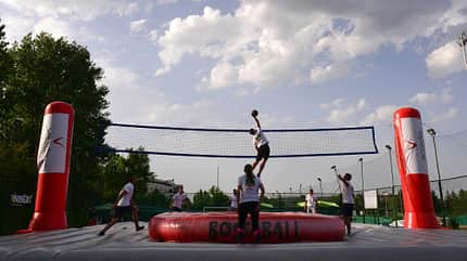 bossaball Eine Gruppe Männer und Frauen befinden sich auf einem aufblasbaren Spielfeld mit einem Netz in der Mitte. Eine Person springt und versucht, an den Ball zu gelangen.